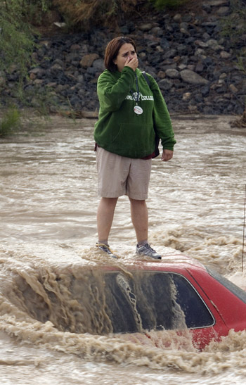 A person standing on the roof of their car that has been overtaken by flood waters. 